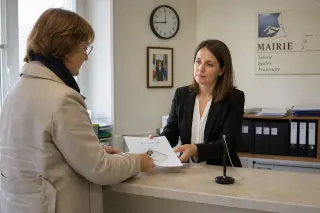 A municipal worker and a visitor exchange documents across a mairie desk, with identification and paperwork visible, in a well-lit, neutral-tone French administrative office